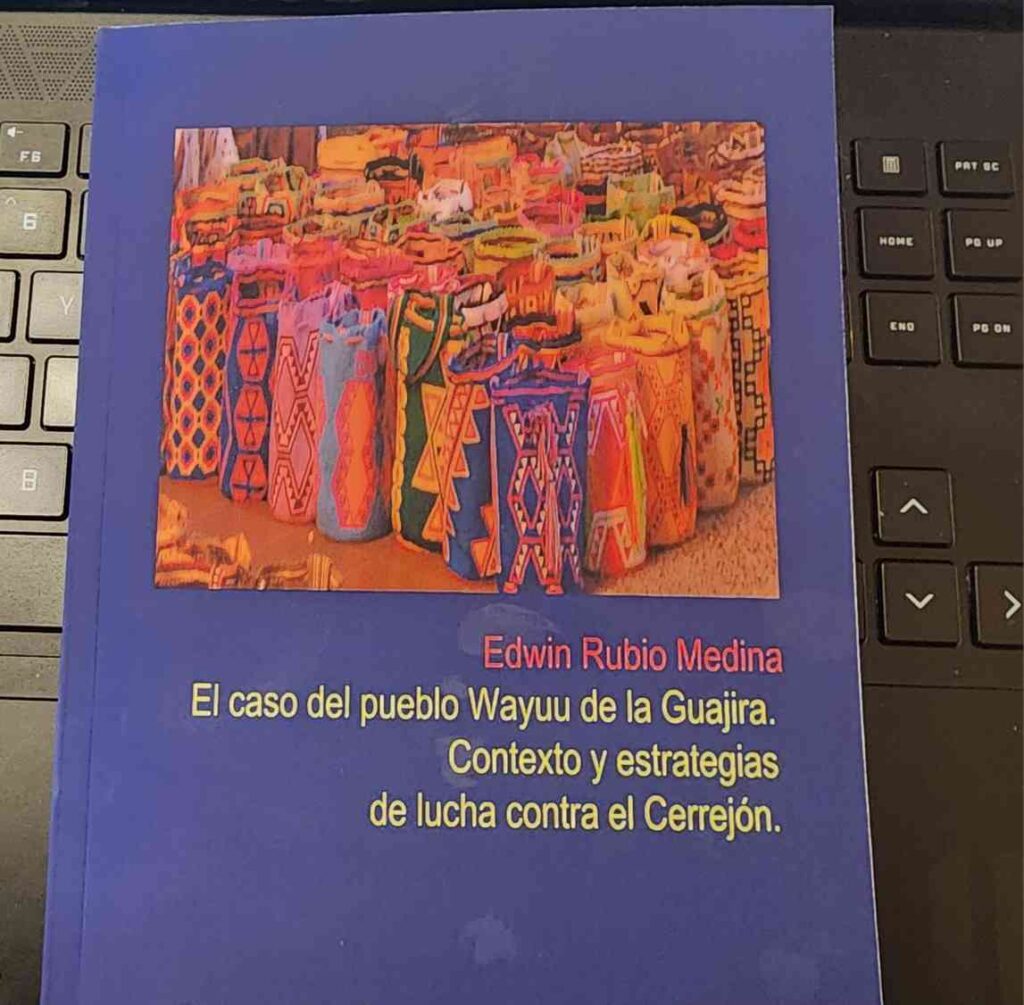 A thin blue book on top of a laptop keykoard. The book has a picture of Wayuu mochilas. Text on the book cover reads "Edwin Rubio Medina" and under that, "The Case of the Wayuu People of the Guajira: Context and Strategies of Struggle against the Cerrejón"